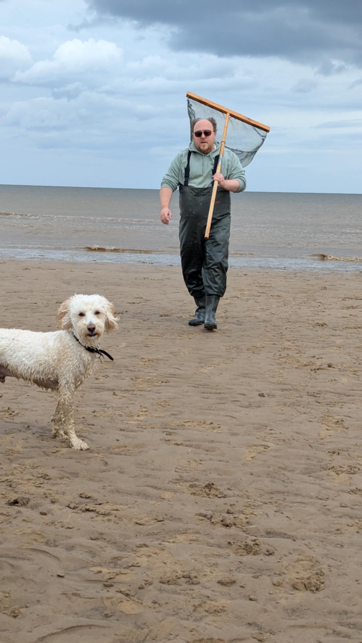 me in waders carrying a shrimp net walking along a sandy beach with a Jacks the dog at low tide